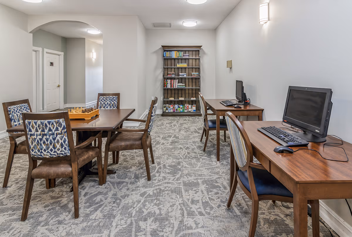 A well-lit room with a patterned carpet featuring a wooden table with four chairs, a chessboard on the table, two desks with computers and chairs, and a bookshelf filled with books and games against the back wall.