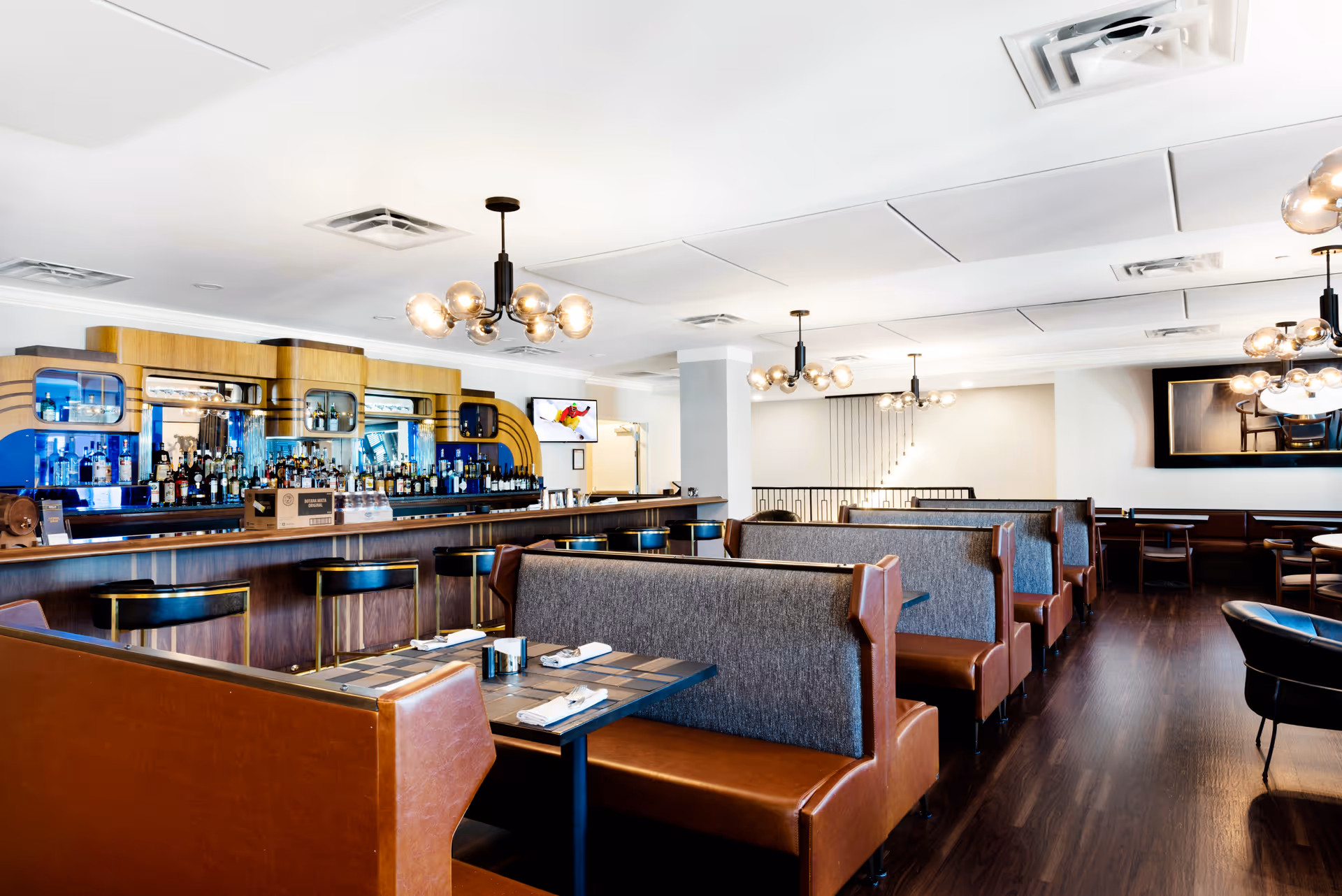Interior view of a modern dining area with brown leather booth seating and dark wood tables set with napkins and utensils. A bar with various bottles and a mirrored back is visible on the left side. The room has contemporary light fixtures hanging from the ceiling and dark wood flooring.