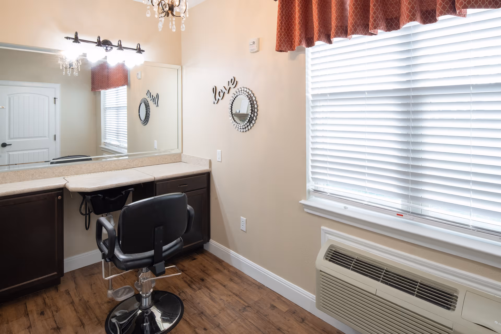 Interior view of a small salon or grooming area with a black salon chair in front of a long countertop with cabinets underneath. A large mirror with lights above it is mounted on the wall. There is a window with closed blinds and a red valance, and a wall-mounted air conditioning unit below the window. The wall has decorative elements including a round mirror and the word 'love'.