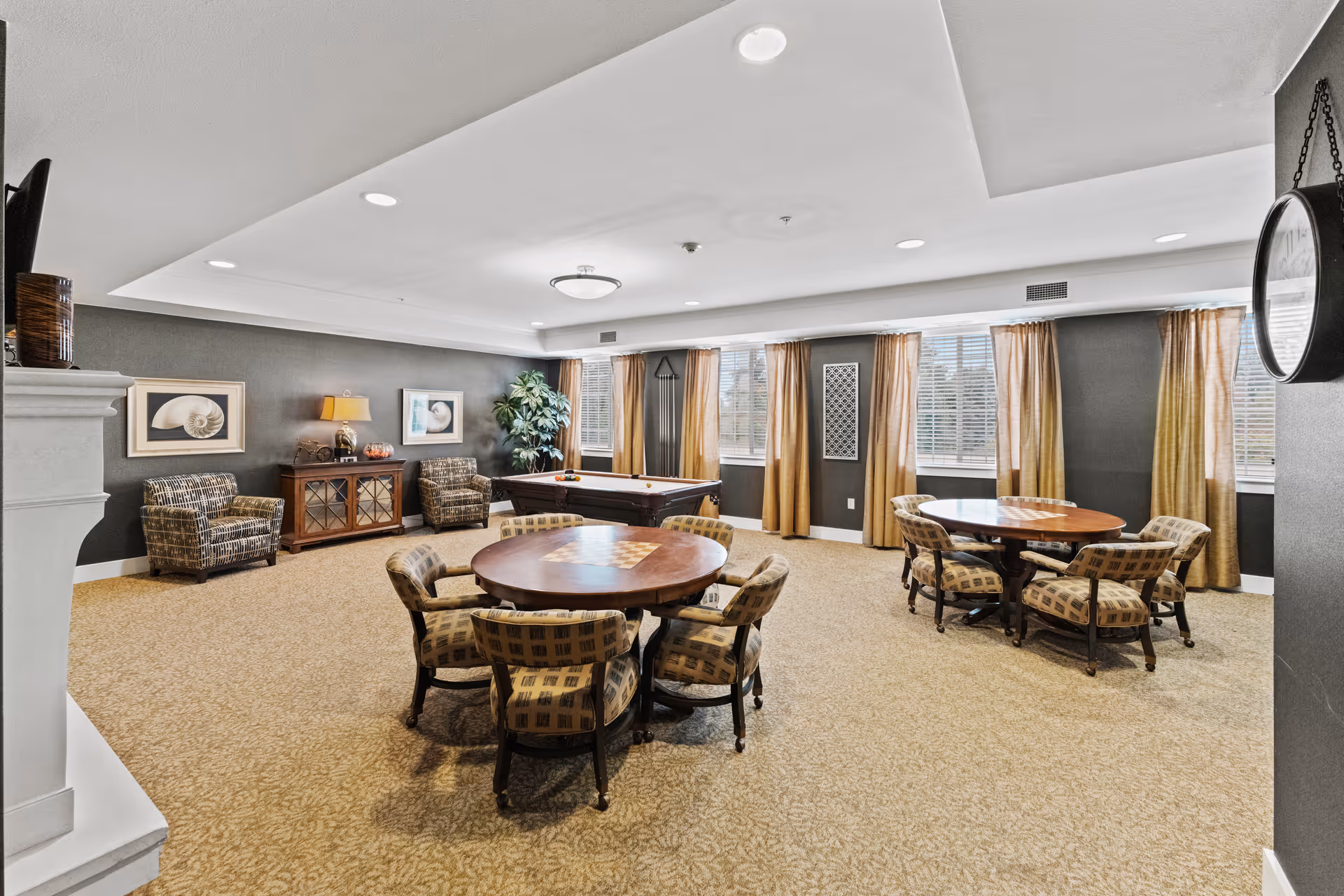 A spacious senior living common room with two round wooden tables each surrounded by six cushioned chairs. The room features beige carpet, gray walls, large windows with yellow curtains, a pool table in the center, two patterned armchairs, a wooden cabinet with a lamp and decorative items, and a large potted plant in the corner.