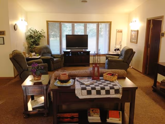 A cozy living room with a large window featuring blinds, a flat-screen TV on a wooden stand, three recliner chairs arranged around the TV, a sofa with a checkered runner on the back table, decorative items including a basket, candle, and flowers, and a carpeted floor.