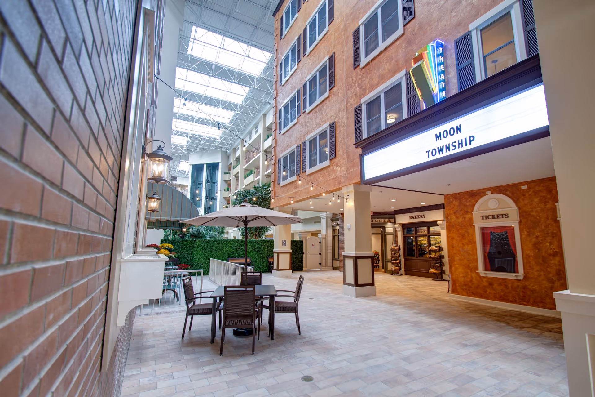 Indoor common area of Tapestry Senior Living Moon Township featuring a high ceiling with skylights, string lights, a table with chairs and an umbrella, a bakery storefront, and a theater marquee sign that reads 'MOON TOWNSHIP'.