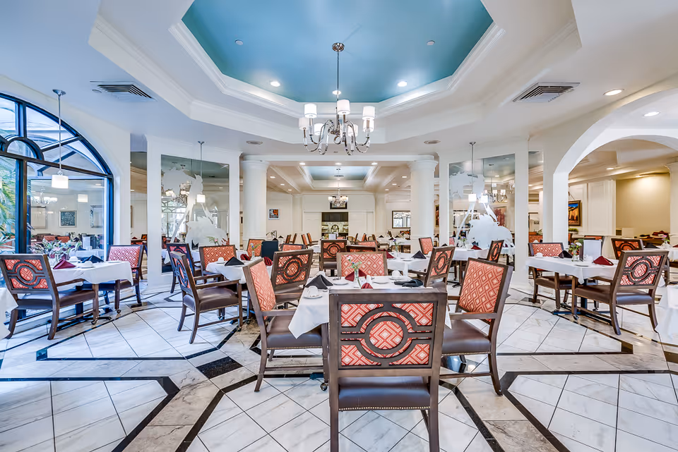 Spacious elegant dining room with multiple set tables and patterned chairs under a decorative coffered ceiling.