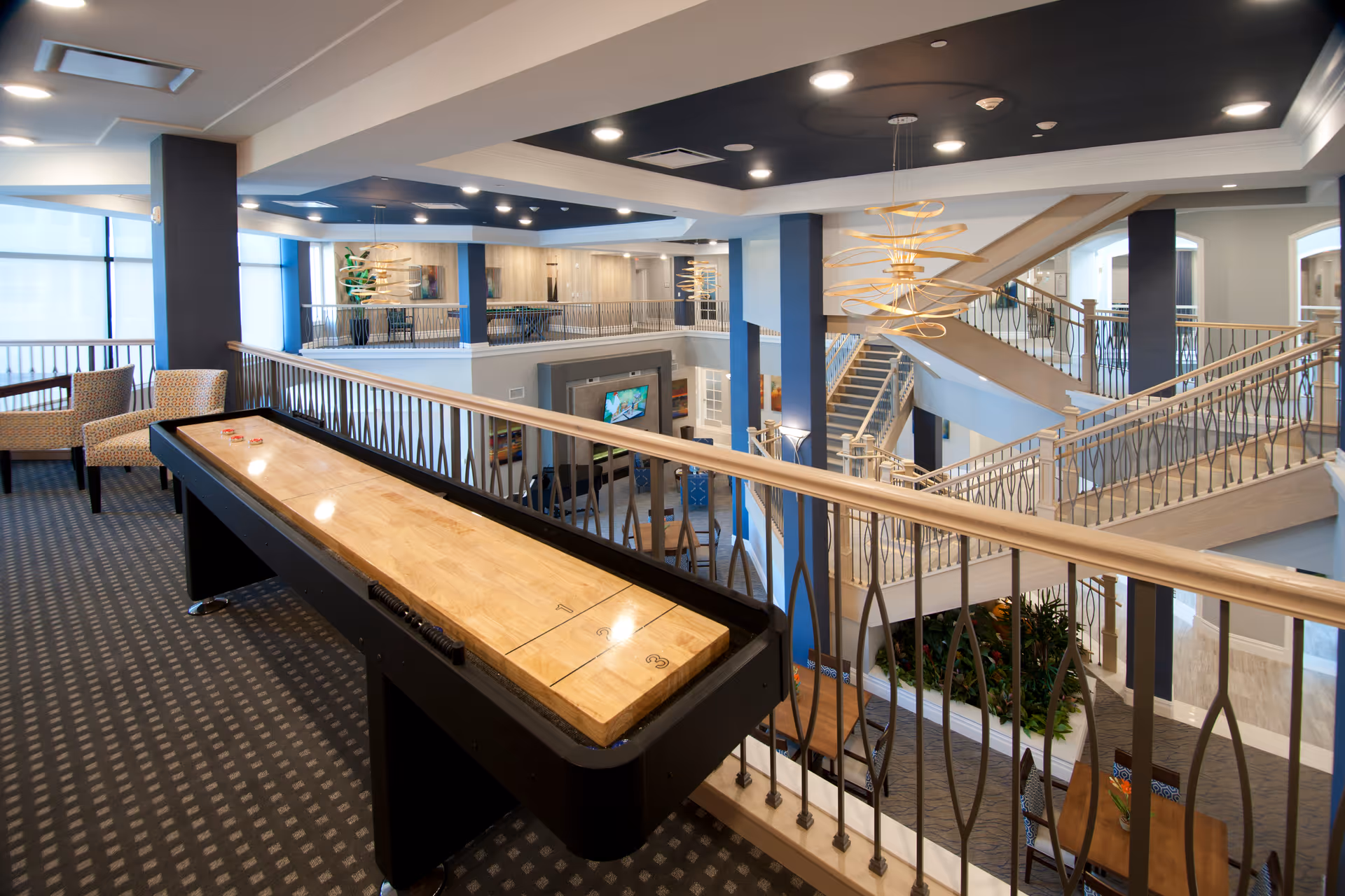 Interior view of a senior living facility featuring a shuffleboard table in the foreground, seating area with chairs to the left, and a multi-level open space with staircases, railings, and modern lighting fixtures. The area is well-lit with natural light from large windows and has a contemporary design with blue and neutral tones.