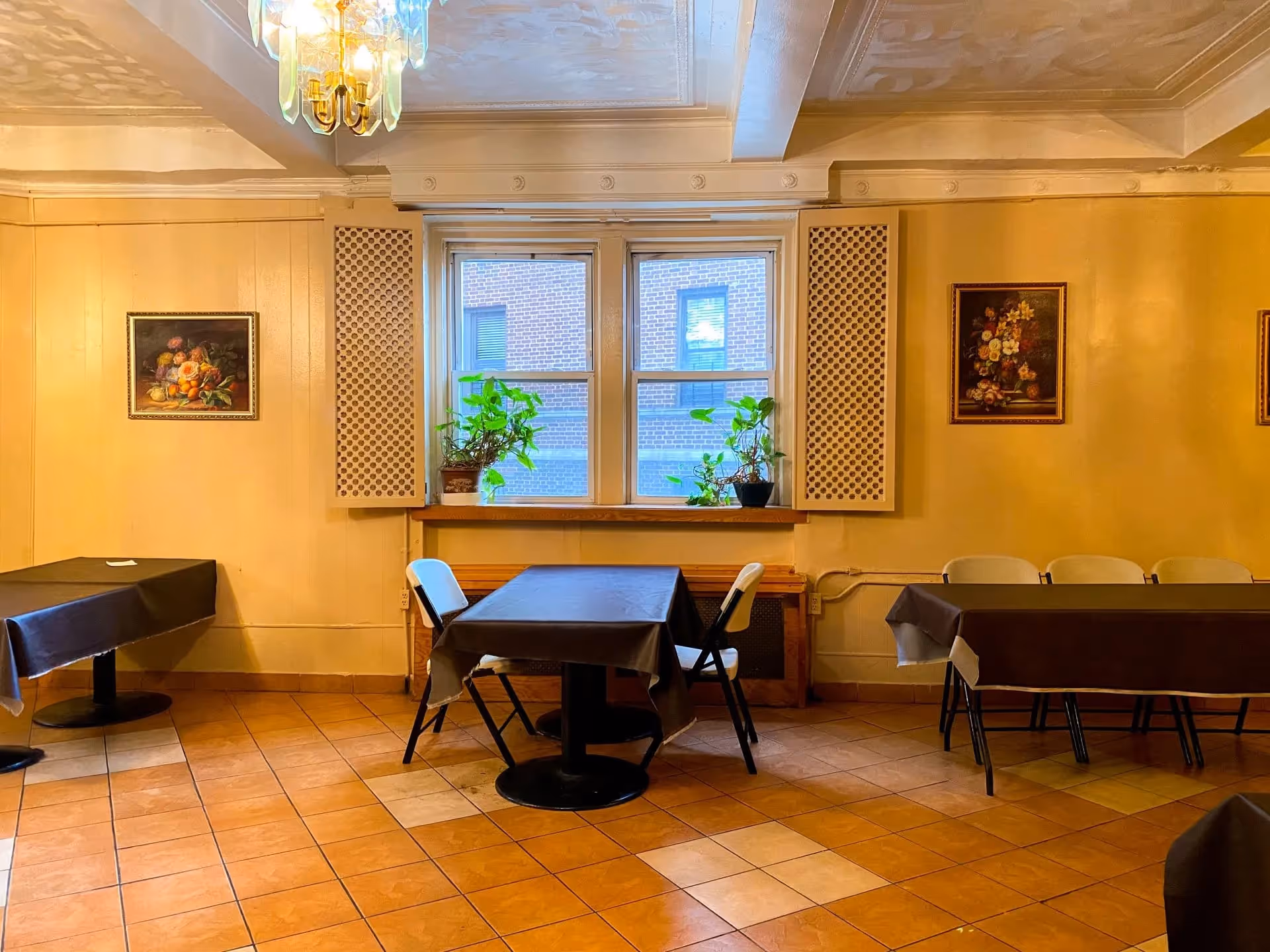 A dining room with several tables covered in dark tablecloths and white folding chairs around them. There are two windows with potted plants on the windowsill, and framed floral paintings on the yellow walls. The floor is tiled in a pattern of light and dark brown squares, and a chandelier hangs from the ceiling.