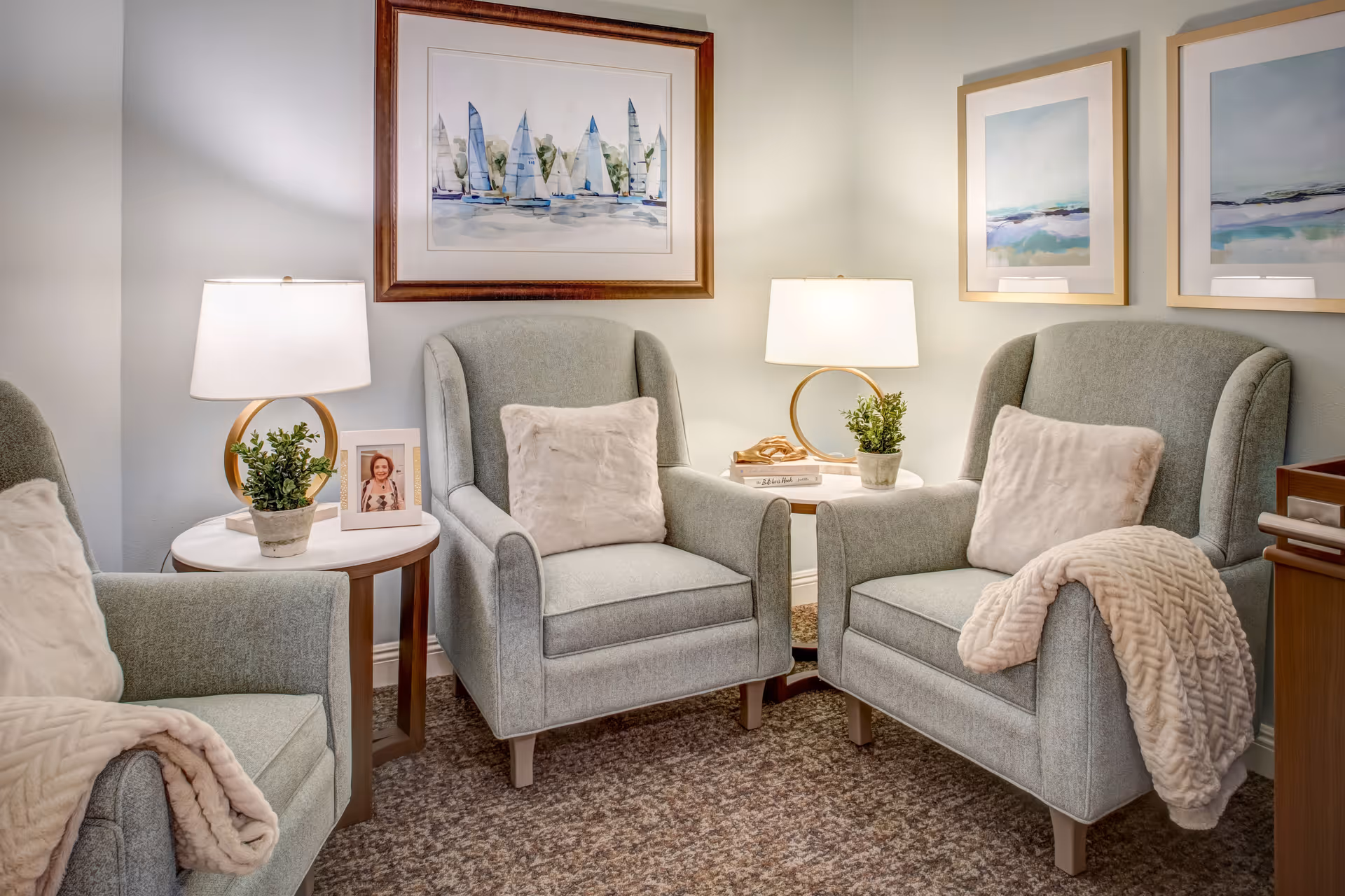 Three light-gray upholstered armchairs arranged around side tables with lamps, framed artwork, throw pillows and blankets in a cozy lounge area.