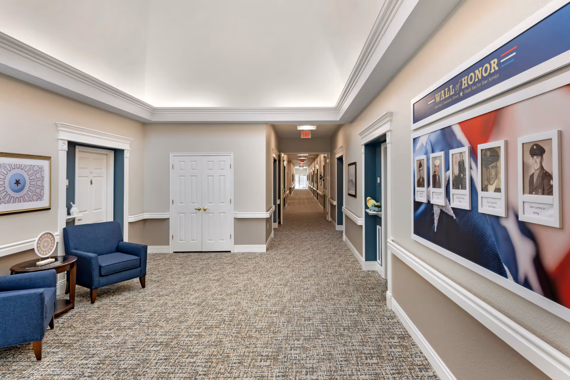 A long, well-lit hallway in a senior living facility with beige walls and carpeted floor. On the right wall, there is a 'Wall of Honor' display featuring framed photos of veterans. Two blue armchairs and a small round table are placed on the left side near a doorway. The hallway extends into the distance with multiple doors and framed pictures along the walls.