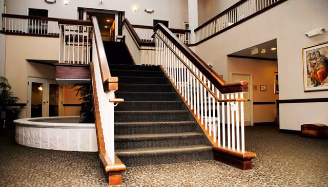 Interior view of a senior living facility lobby featuring a wide staircase with dark carpet and wooden handrails leading to an upper floor. The area has patterned carpet flooring, white walls, and a railing overlooking the lobby from the second floor. There is a circular white structure to the left, possibly a planter or seating area, and framed artwork on the walls.