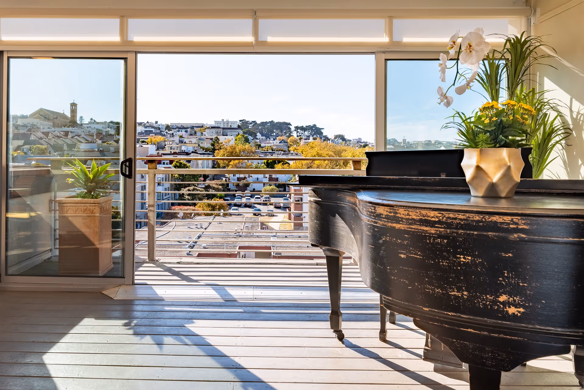 Sunlit interior room with a worn black grand piano by sliding glass doors opening to a balcony and city view.