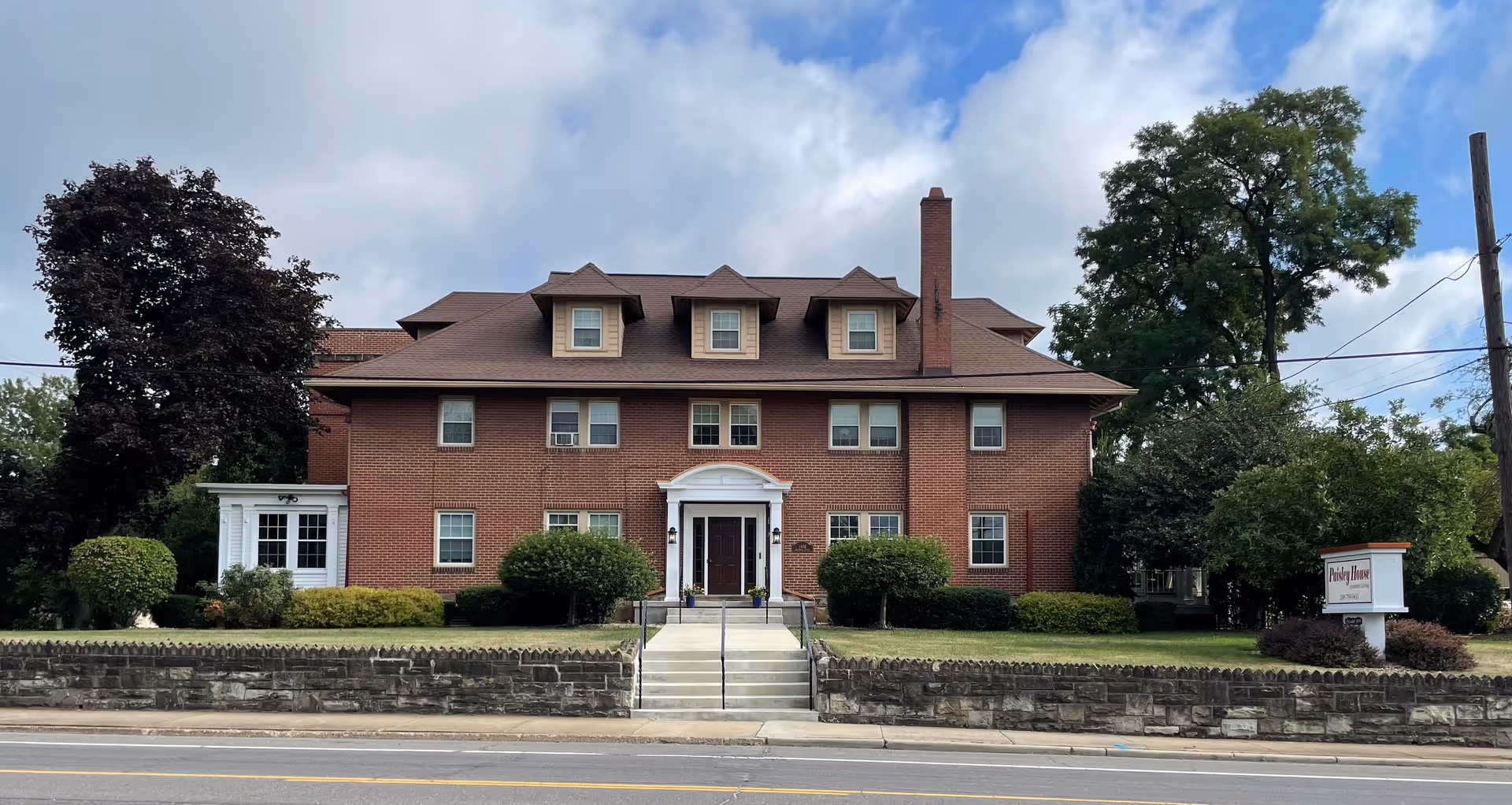 Front exterior view of a large, three-story brick building with a brown roof and multiple windows. There are bushes and trees surrounding the building, a stone wall along the sidewalk, and a sign on the right side that reads 'Paisley House'. The sky is partly cloudy.