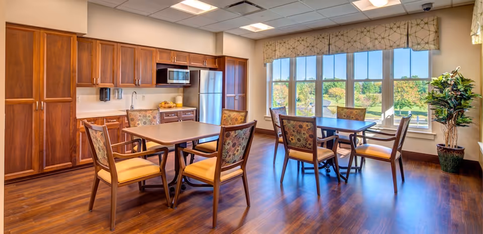 A bright dining area with two tables and eight chairs featuring floral upholstery and yellow cushions. The room has large windows with a scenic view of greenery outside, wooden flooring, and a kitchen area with wooden cabinets, a stainless steel refrigerator, and a microwave.