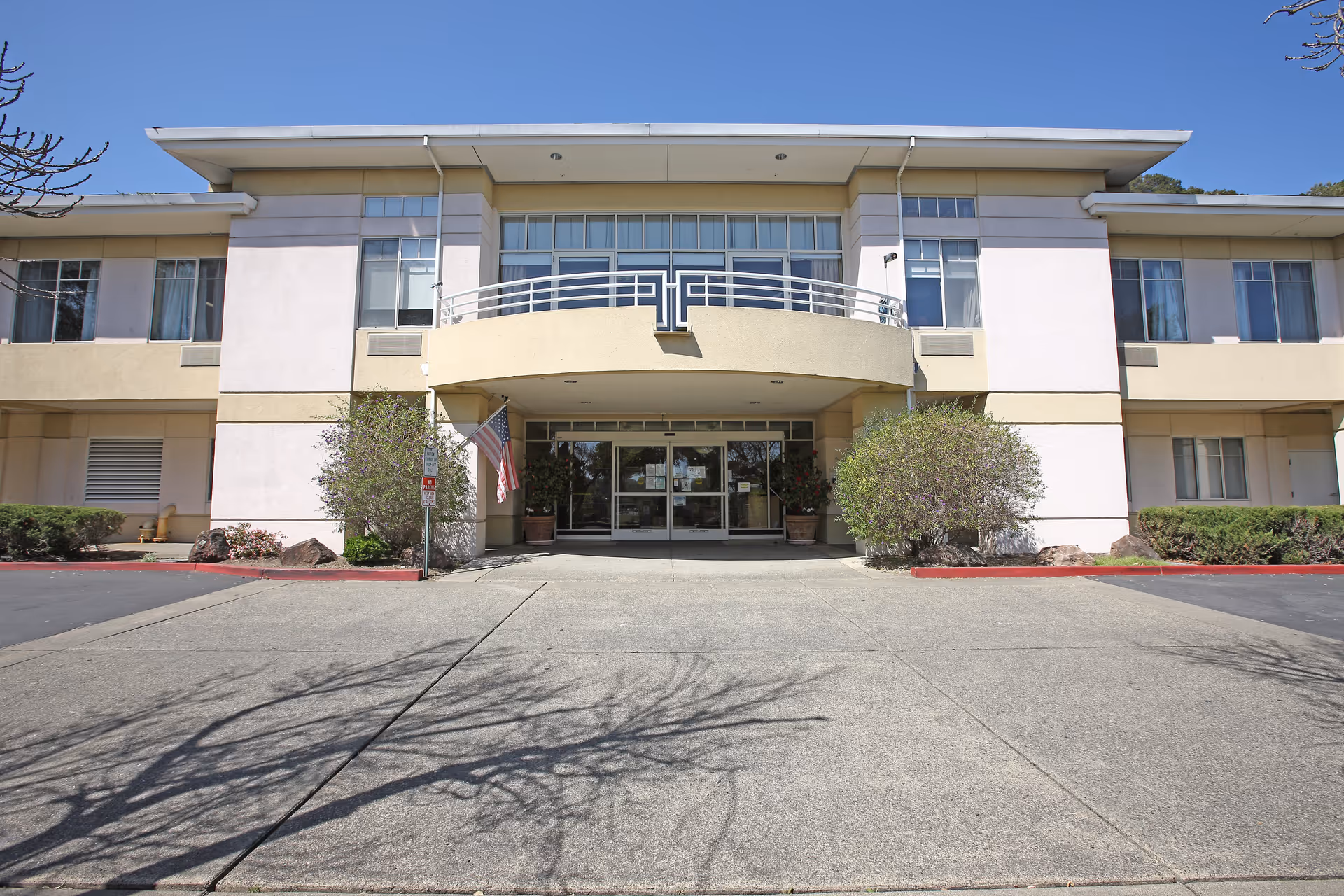 Front exterior view of Smith Ranch Skilled Nursing & Rehabilitation Center building with a wide concrete driveway, two bushes flanking the entrance, an American flag on the left side, and clear blue sky above.