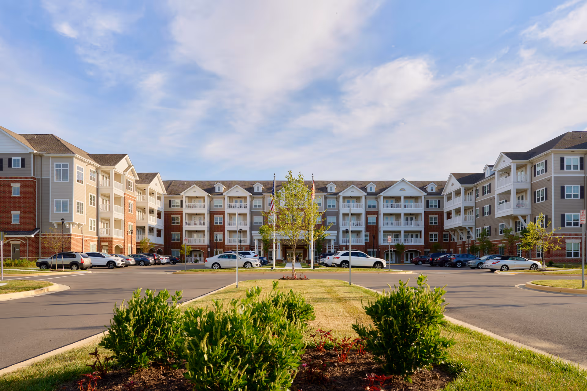 Front exterior view of a large senior living facility named Harmony at Enterprise, featuring a multi-story building with balconies, a parking lot with several cars, and landscaped greenery in the foreground under a partly cloudy sky.