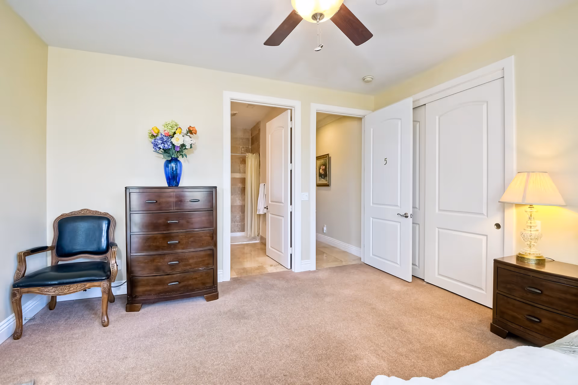 Bright bedroom featuring a wooden dresser topped with a vase of flowers, an upholstered chair, open doors to a bathroom and closet, and a bedside lamp.
