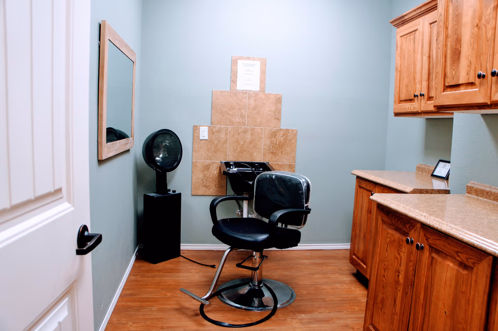 A small salon room with a black salon chair in front of a hair washing station. The room has light green walls, wooden cabinets with granite countertops on the right, a mirror on the left wall, and a hair dryer standing on a black pedestal next to the chair.