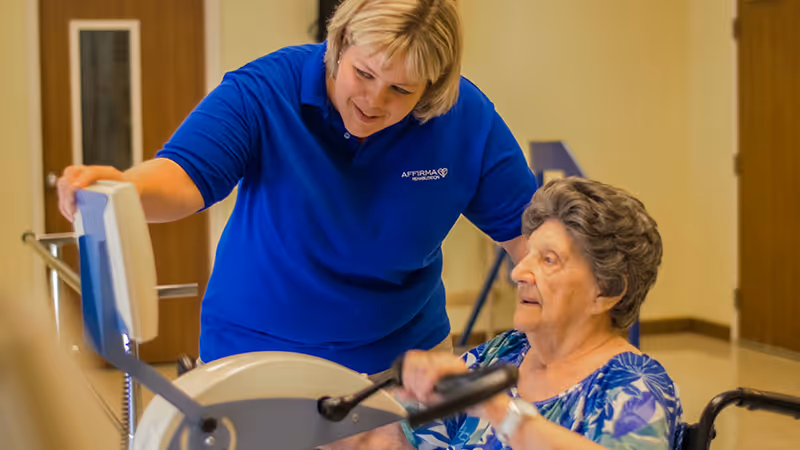 A caregiver in a blue shirt assists an elderly woman in a wheelchair using an exercise machine in an indoor rehabilitation or healthcare facility.