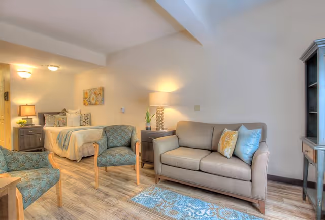 A cozy and well-lit senior living room and bedroom area featuring a beige sofa with decorative pillows, two patterned armchairs, a small blue and beige rug, a wooden side table with a lamp, and a bed with multiple pillows and a floral painting above it. The flooring is wood-style, and the walls are painted white.