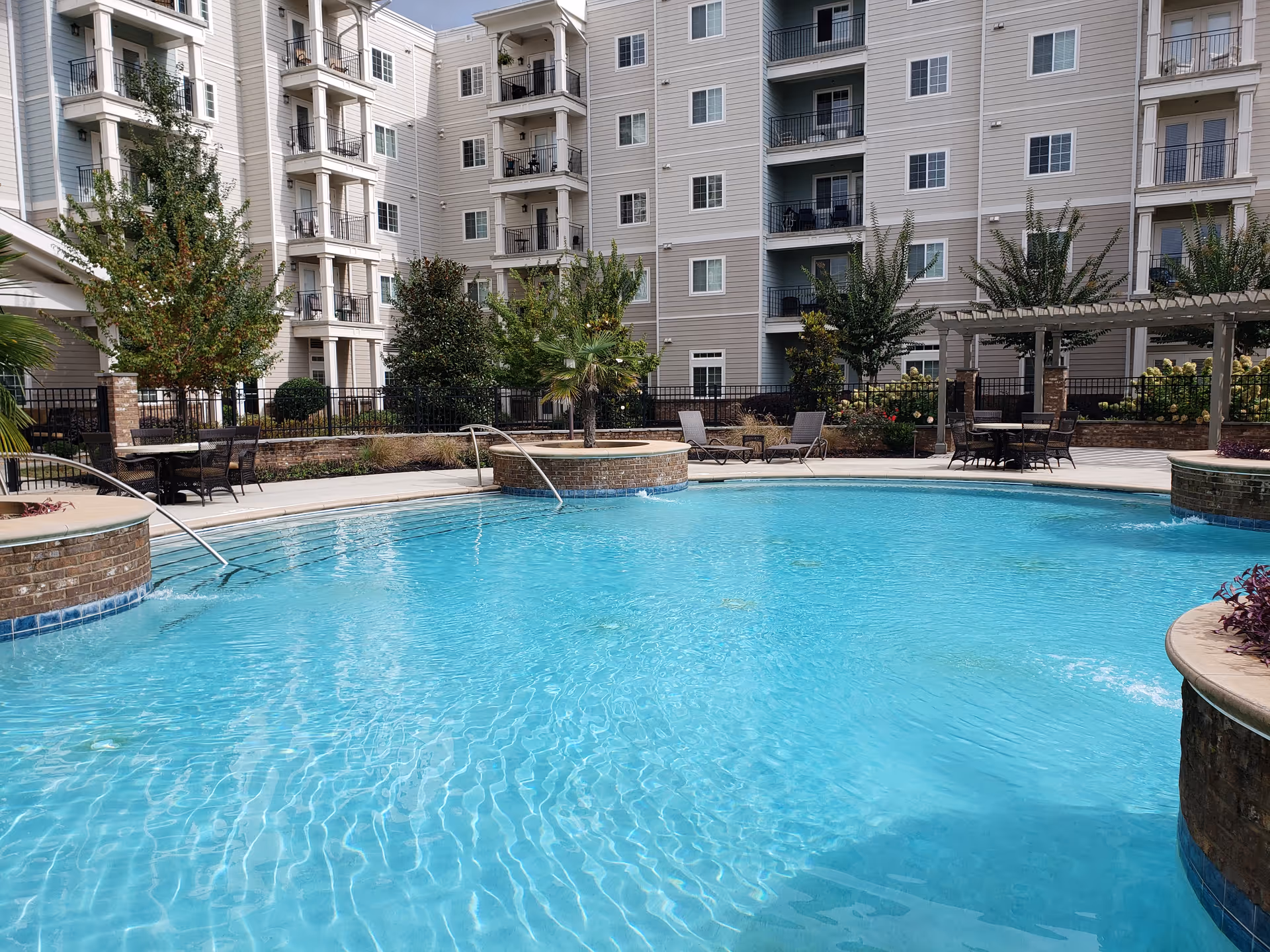 Outdoor swimming pool with clear blue water surrounded by a patio area with tables, chairs, and lounge chairs. The pool area is landscaped with trees and plants, and a multi-story residential building is visible in the background.
