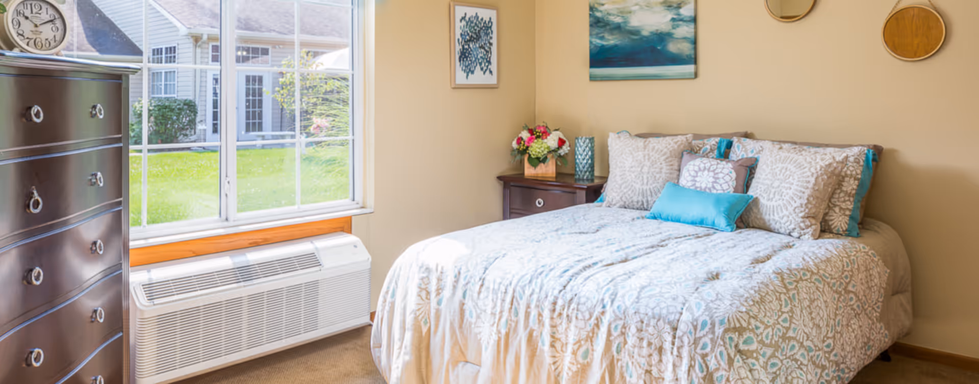 Sunny bedroom with a made bed with decorative pillows, a nightstand with flowers, a dresser, and a large window overlooking a courtyard.