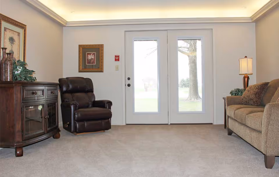 A cozy living room area with a brown leather recliner chair on the left, a wooden cabinet with decorative vases and a plant, and a beige fabric sofa with patterned cushions on the right. There is a table lamp on a side table next to the sofa. The room has light-colored walls and carpeted floor, with double glass doors in the center leading outside, showing a tree and greenery through the glass.
