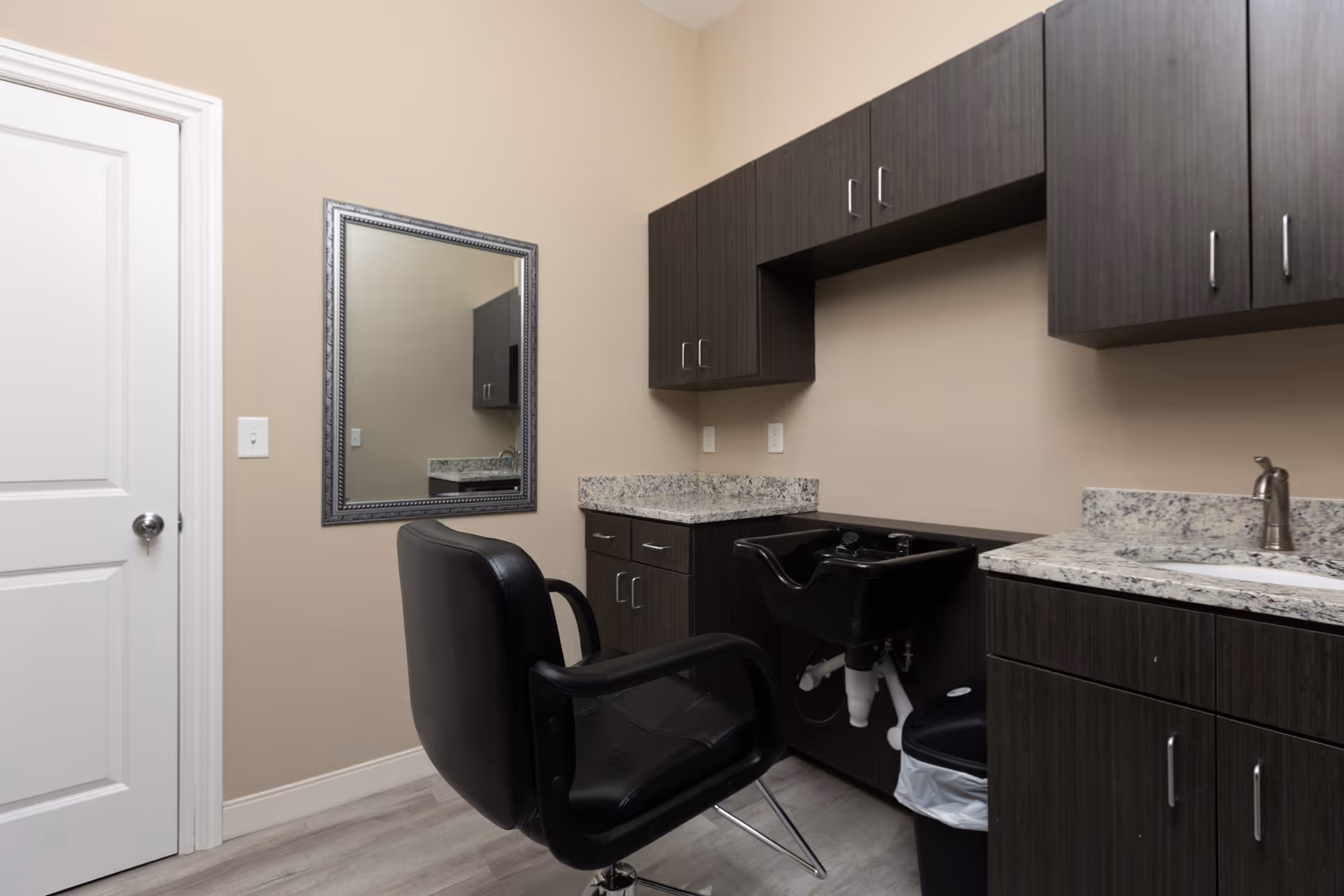 Interior view of a small salon area in an assisted living facility featuring a black salon chair in front of a black wash basin. The room has dark wood cabinets with granite countertops, a wall-mounted mirror, and a white door on the left side.