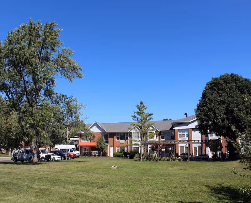 Two-story senior living facility building with a large grassy front lawn, trees, and parked cars under a clear blue sky.