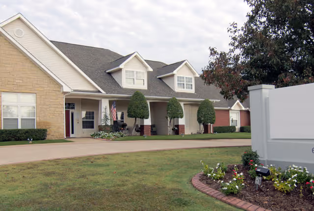 Exterior view of a single-story residential building with a stone and brick facade, multiple windows, and a driveway. There are neatly trimmed trees and a small garden bed with flowers near a blank white sign in the foreground.