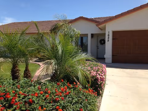 Exterior view of a single-story senior living home with palm trees and colorful flowers.