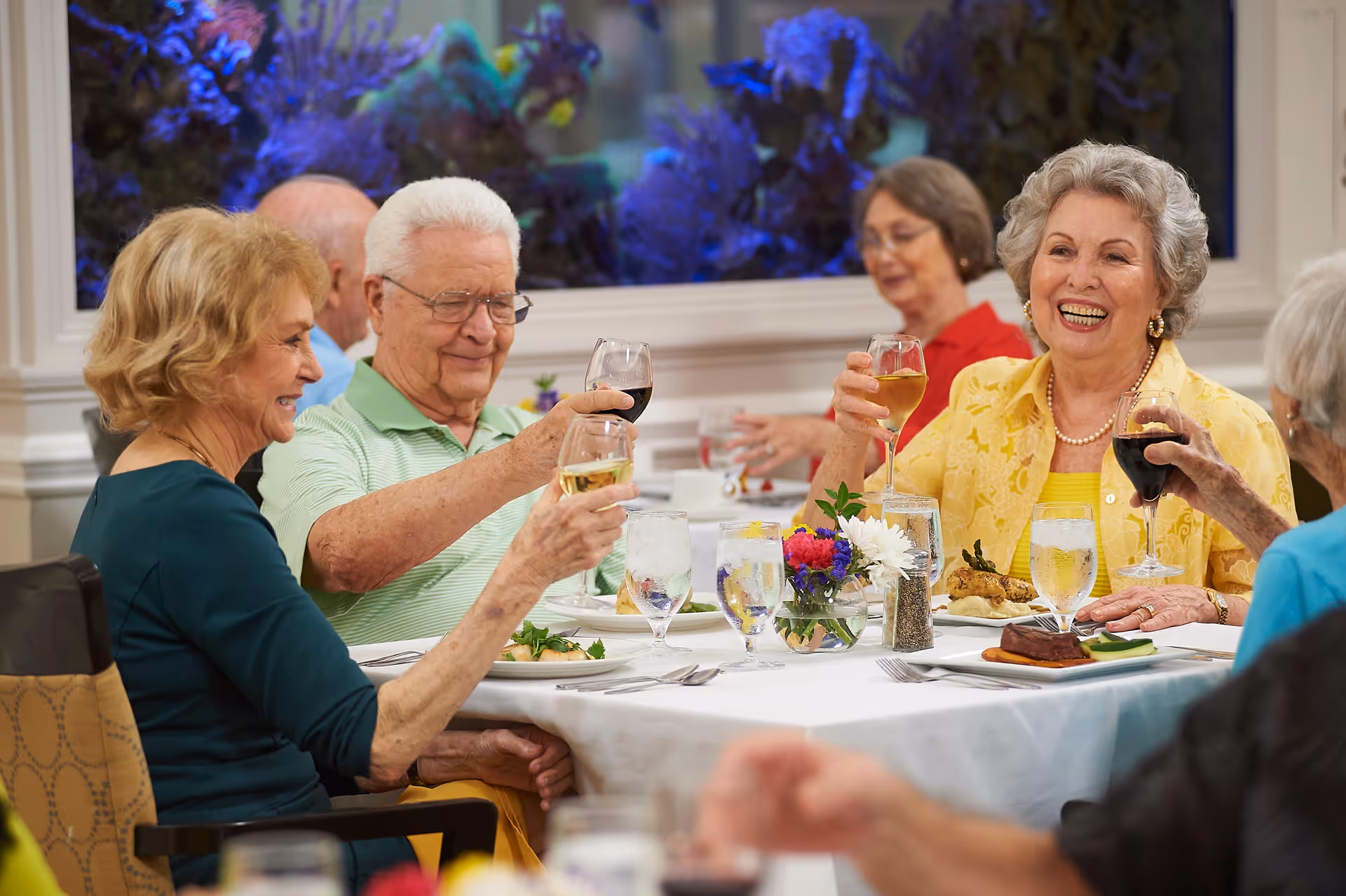 A group of elderly people sitting around a dining table in a well-lit room, raising their glasses in a toast and smiling. The table is set with plates of food, glasses of water, and a small flower arrangement in the center. The background features a large window with a blue-lit aquarium.