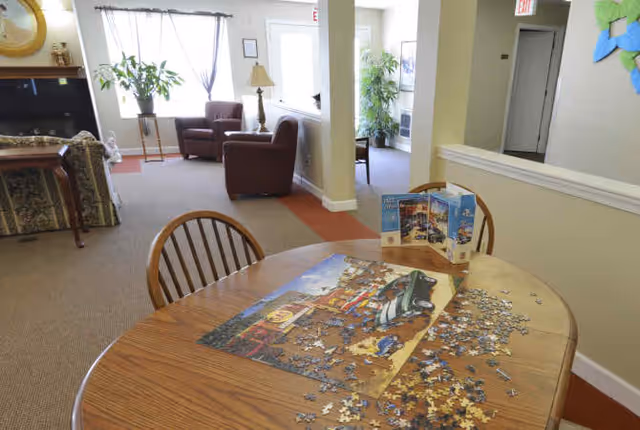 A cozy common area with a wooden table in the foreground partially covered with a jigsaw puzzle and puzzle box. In the background, there are two brown armchairs, a lamp, a plant near a window with sheer curtains, and a fireplace with a mirror above it. The room has beige walls and carpeted floors with a red border.