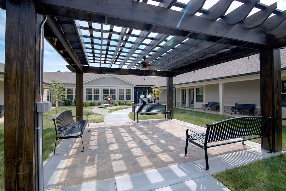 Outdoor seating area with black metal benches under a wooden pergola casting shadows on the paved ground. Surrounding the area is a grassy courtyard with a pathway leading to a building with multiple windows and doors.