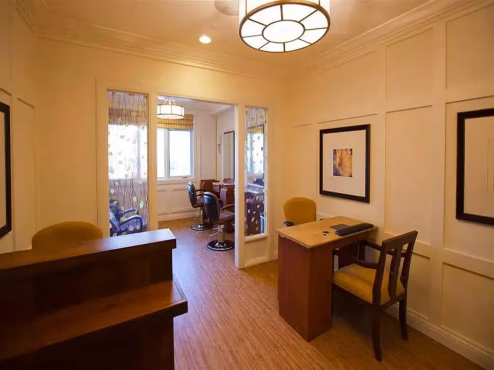 Interior view of a well-lit room with wooden flooring and beige walls. The room features a small desk with two chairs, a framed picture on the wall, and a reception counter. Through an open doorway, another room is visible with salon-style chairs and mirrors, suggesting a hair salon or grooming area.