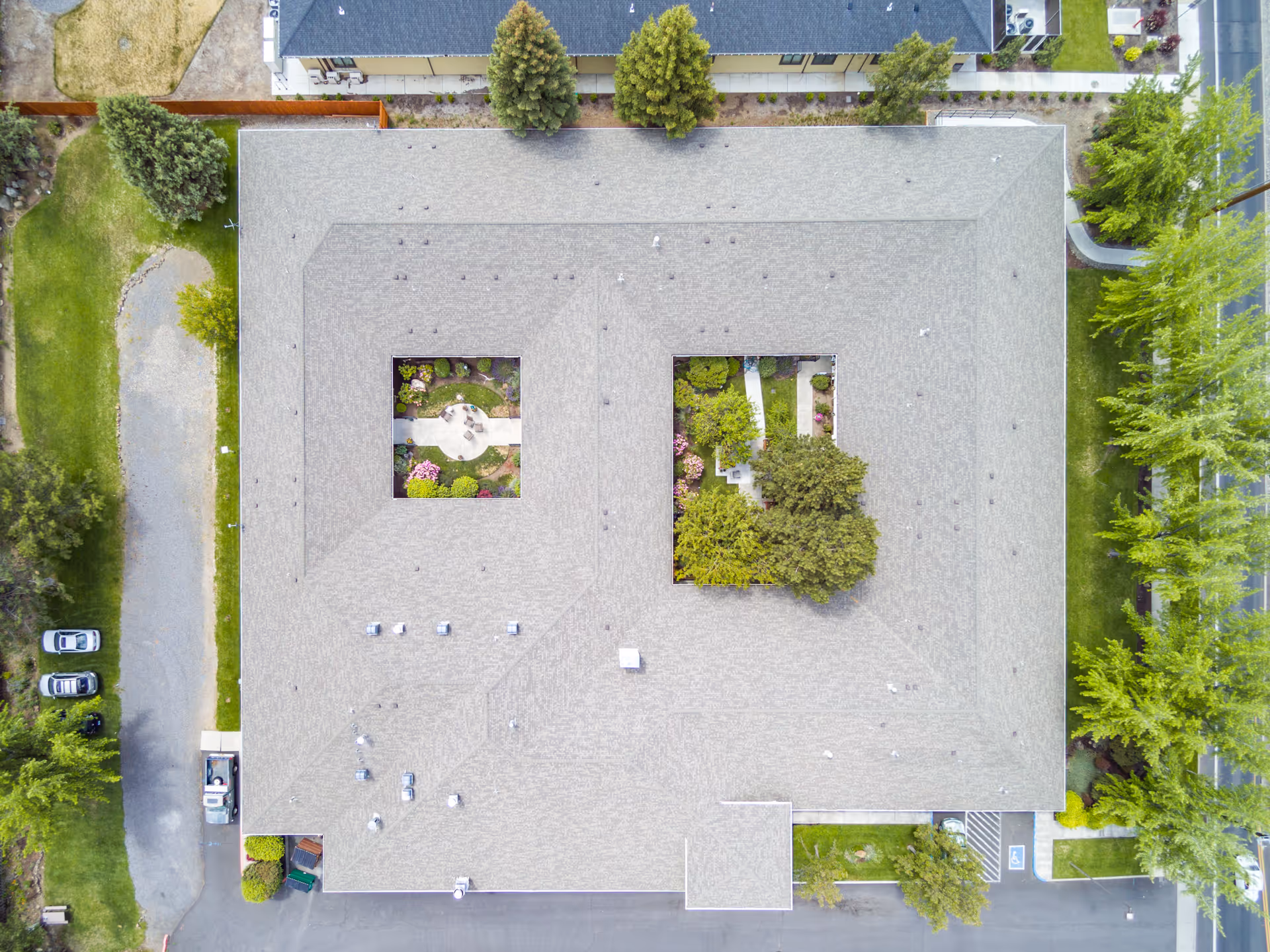 Aerial view of a large rectangular building with two open courtyard gardens in the center, surrounded by trees, parking spaces, and pathways.