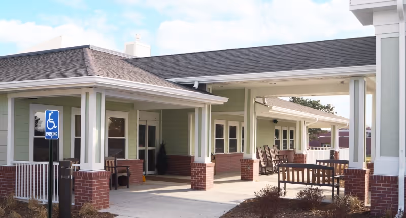 Covered front entrance of a care facility with benches and rocking chairs under a porch and a handicap parking sign.