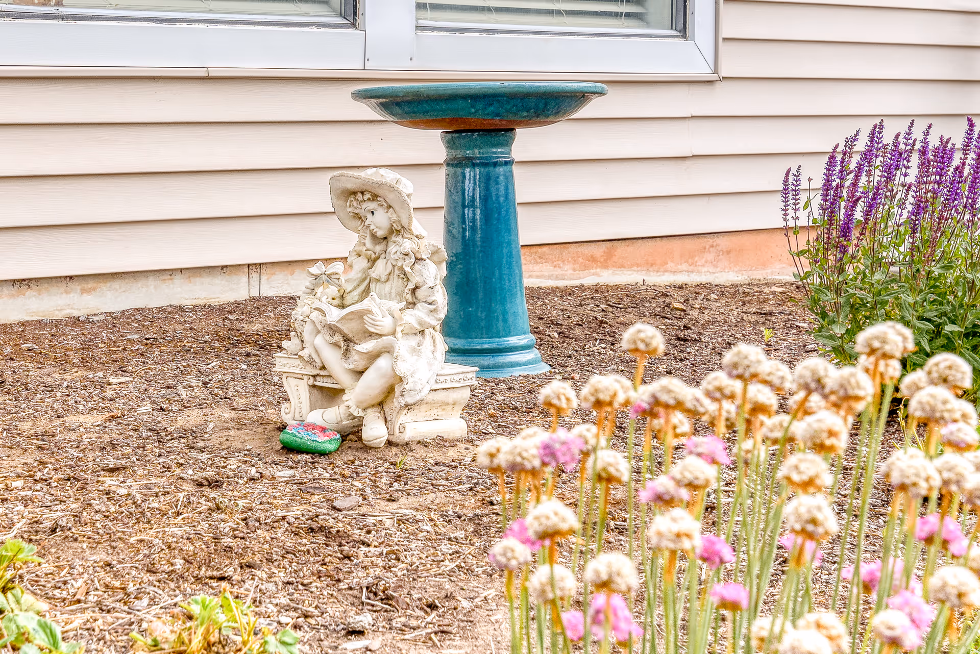 Garden bed with a small statue of a girl reading, a teal birdbath, and pink and white flowers in front of beige siding.
