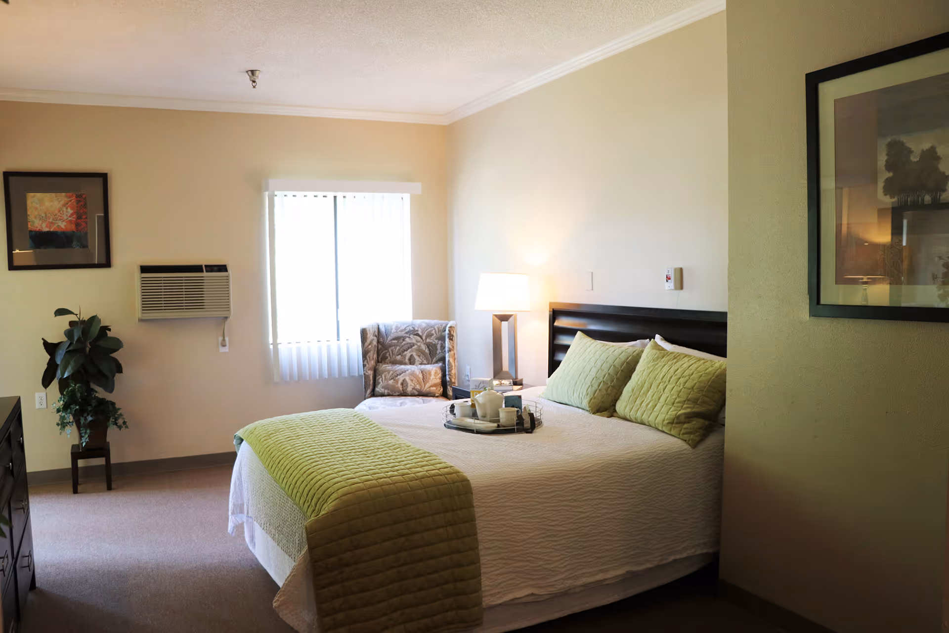 A cozy bedroom with a neatly made bed featuring a white quilt and green pillows and throw blanket. A tray with a teapot and cups is placed on the bed. There is a patterned armchair next to a window with vertical blinds, a lamp on a bedside table, a framed picture on the wall, an air conditioning unit, and a potted plant on a small stand.