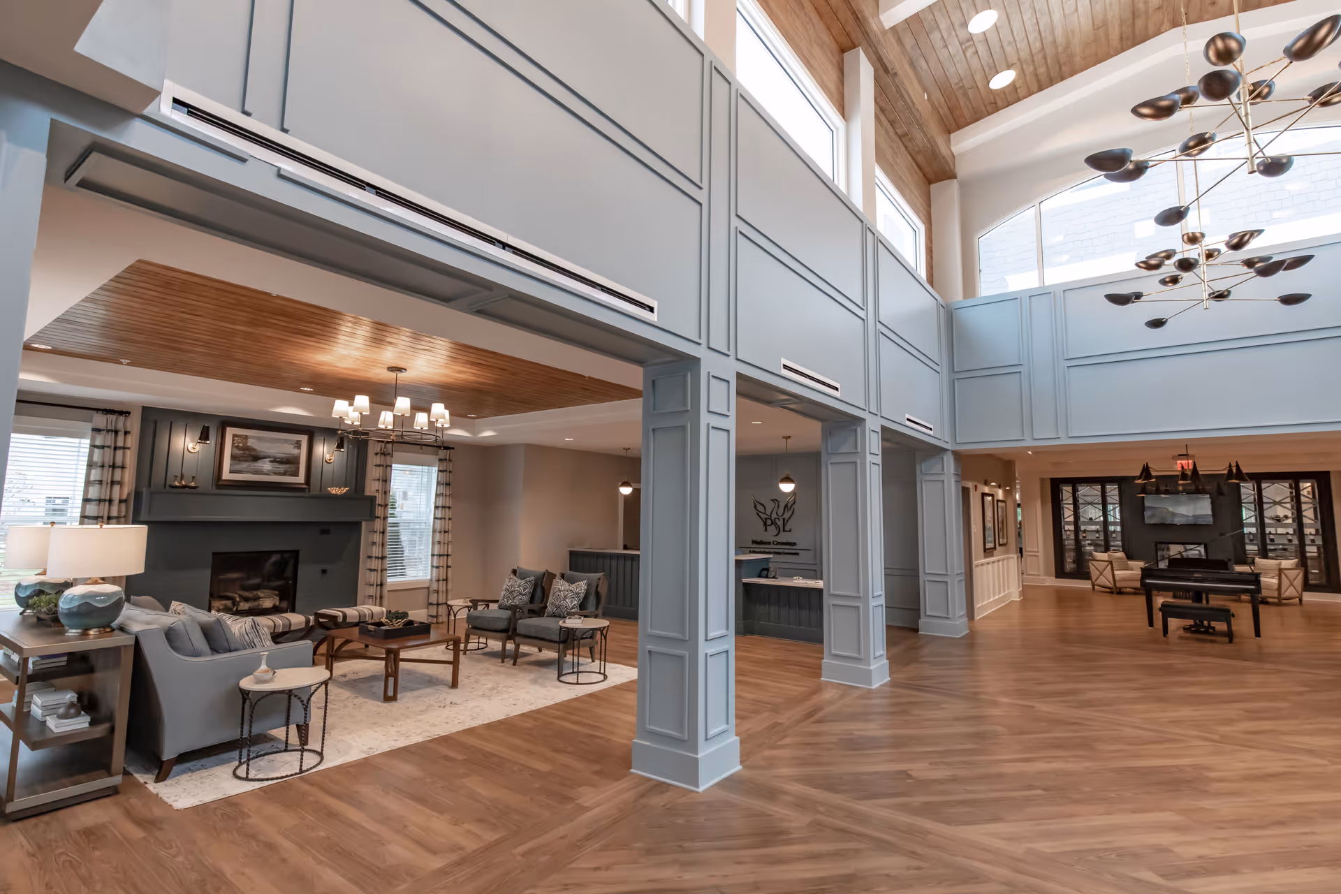 Spacious and well-lit senior living facility common area with high ceilings, large windows, and wooden flooring. The room features a seating area with sofas, armchairs, a coffee table, and a fireplace. There is a reception desk in the background and a piano with additional seating further back. Modern chandeliers hang from the ceiling, and the walls are painted in soft neutral tones.