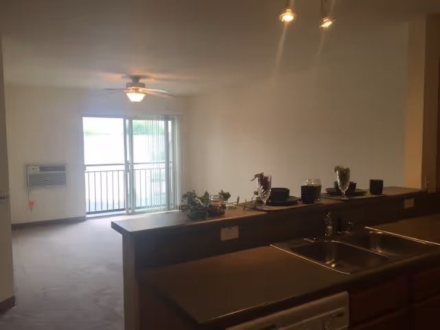 Interior view of a senior living apartment at Berkshire-Oconomowoc showing a kitchen counter with a double sink and some decorative items. Beyond the counter is a living area with carpeted floor, a ceiling fan with light, a wall-mounted air conditioner, and sliding glass doors leading to a balcony.