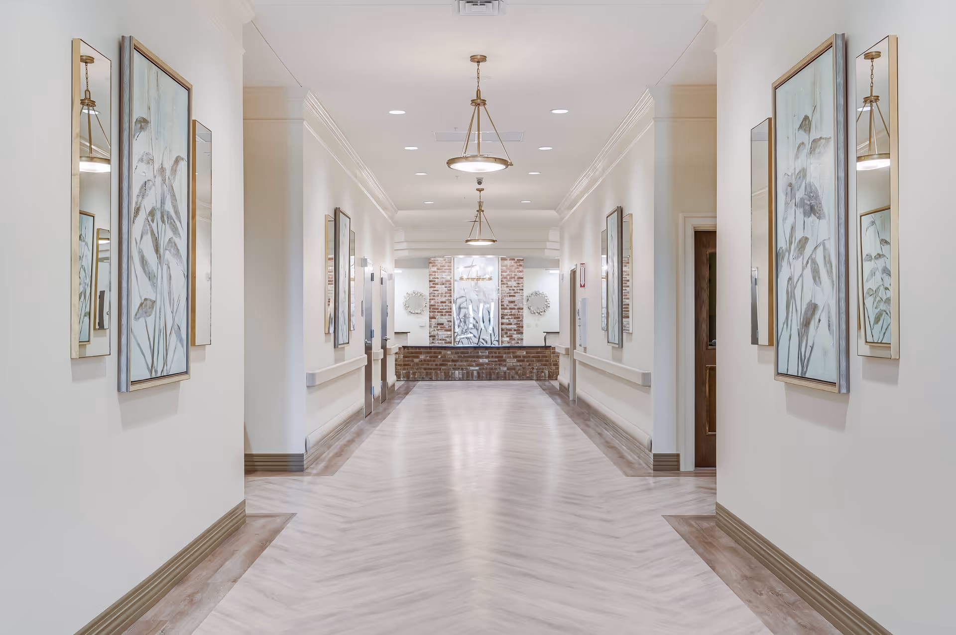 Bright, wide hallway in a senior living facility lined with framed artwork, handrails, and pendant lights leading to a reception area.