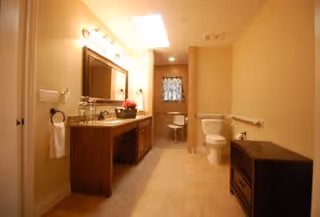 A spacious bathroom with beige walls and tiled floor featuring a large wooden vanity with a sink and mirror, a toilet, a small chair near a window with a patterned curtain, and a dark wooden cabinet on the right side.