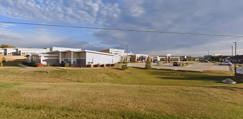 Exterior view of Hawkeye Care Center showing a single-story building with a parking lot and a grassy area in the foreground under a partly cloudy sky.