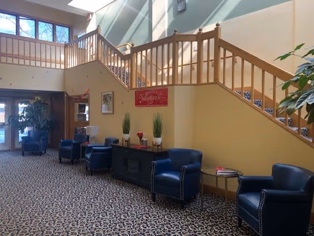 Interior view of a senior living facility lobby area with a staircase leading to an upper floor. The space features patterned carpet, several blue armchairs, a black cabinet with decorative plants and flowers, and a sign on the wall that reads 'Happy Valentine's Day'. Large windows allow natural light to brighten the area.