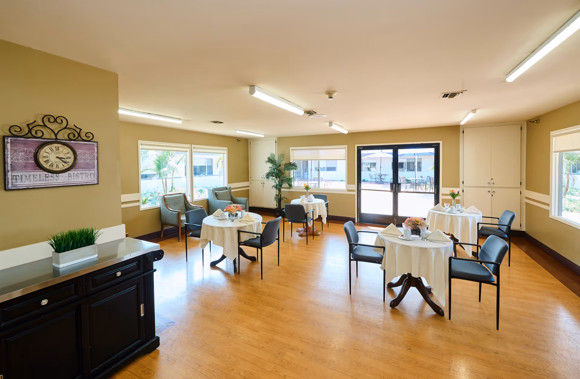 Bright communal dining room with round tables set with tablecloths and chairs, large windows and glass doors overlooking an outdoor patio.
