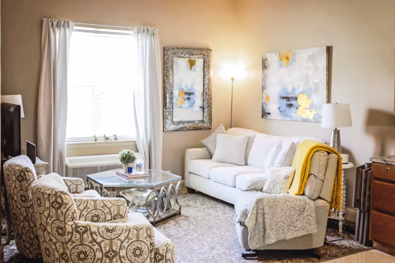 A cozy living room with a white sofa, a patterned armchair, and a recliner covered with a gray throw and a yellow blanket. A glass coffee table with decorative items sits in front of the sofa. The room has a window with white curtains, a floor lamp, a table lamp, and abstract artwork on the walls.