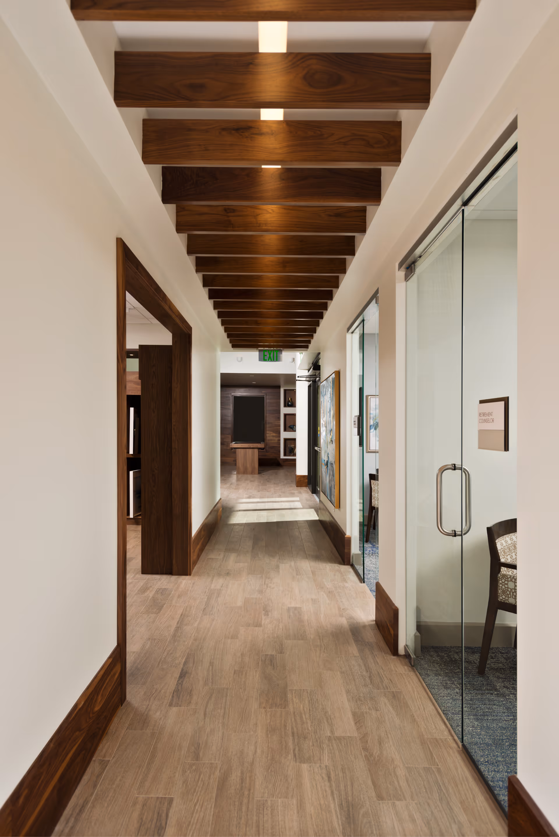 A well-lit hallway with wooden beams on the ceiling and wood trim along the floor edges. The floor is covered with light wood-patterned tiles. On the right side, there is a glass door leading to a room with a chair visible inside. The hallway extends to a room at the end with a dark board and some shelves.