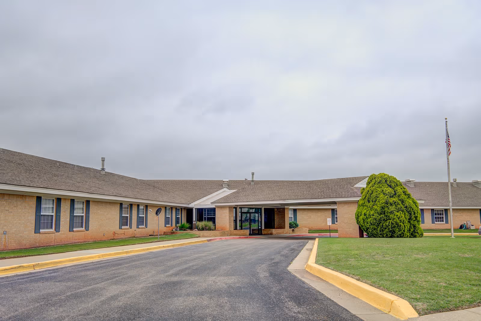Front exterior of a single-story brick assisted living building with a driveway, entrance canopy, flagpole, and manicured lawn under an overcast sky.