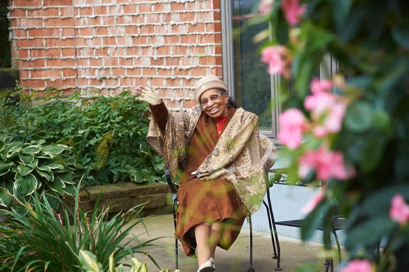 An elderly woman wearing a beige hat, glasses, and a patterned shawl is sitting on a metal chair outdoors, smiling and waving. She is surrounded by green plants and pink flowers, with a brick wall and window in the background.