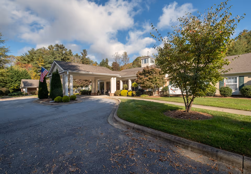 Exterior view of Carolina Reserve of Hendersonville senior living facility showing the main entrance with a covered drop-off area, surrounded by trees, bushes, and a well-maintained lawn under a partly cloudy sky.