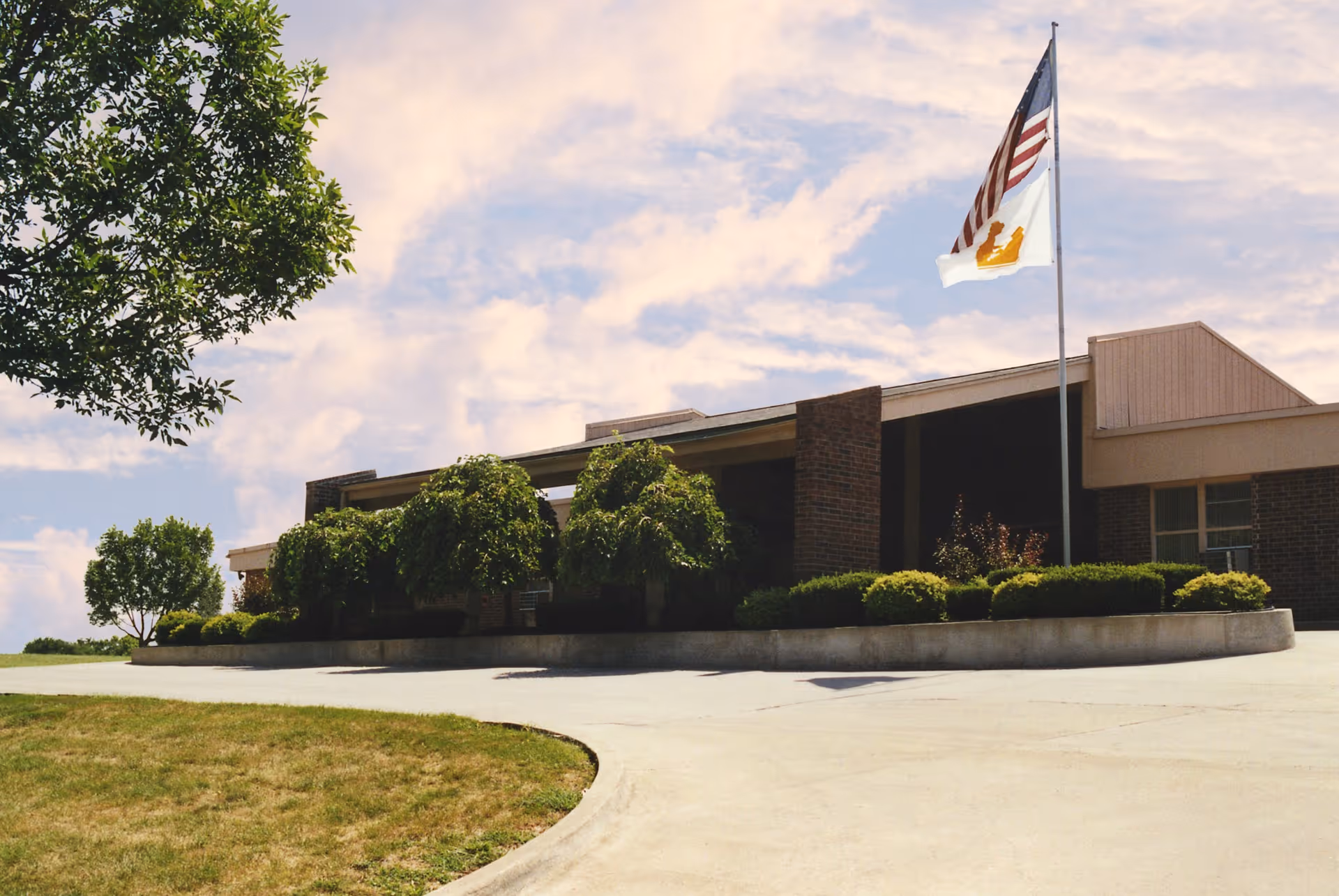 Exterior view of a single-story brick building with a flagpole displaying the American flag and another flag with a crown symbol. The building is surrounded by neatly trimmed bushes and trees under a partly cloudy sky.