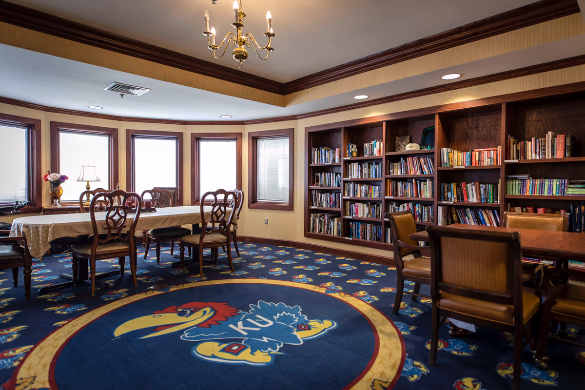 A cozy library room with wooden bookshelves filled with books along one wall. There are several wooden chairs and tables, including a long table covered with a white tablecloth near windows with blinds. The carpet features a large colorful logo of a bird with the letters 'KU' in the center. A brass chandelier hangs from the ceiling.